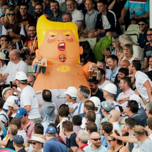 A man in a Donald Trump baby blimp outfit walks back into the Hollies stand carrying beers.